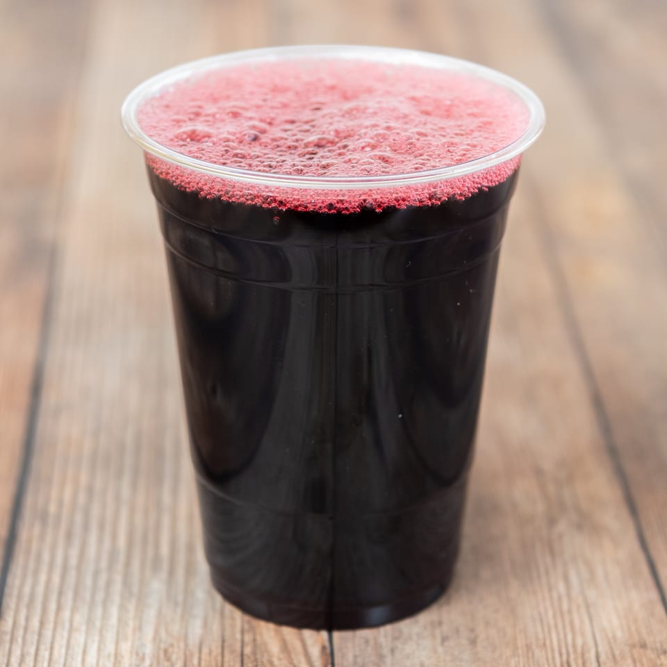 Dark red juice with a foamy top in a clear plastic cup on a wooden surface.