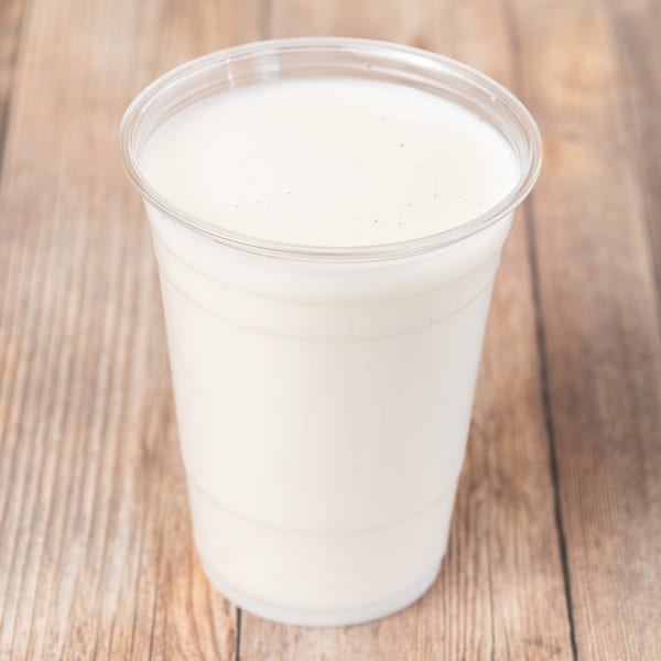 A clear plastic cup filled with a creamy white beverage, possibly a milkshake or smoothie, on a wooden surface.