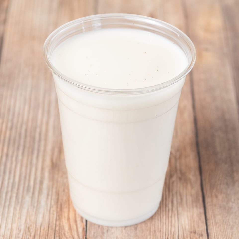 A clear plastic cup filled with a creamy white beverage, possibly a milkshake or smoothie, on a wooden surface.