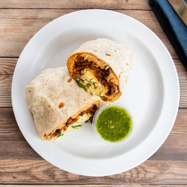 Two halves of a burrito filled with meat, rice, and vegetables, served with green salsa.