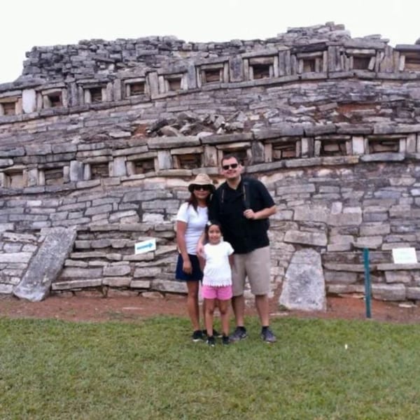Family of three posing in front of a large, ancient stone structure with many small square openings.