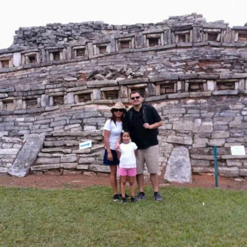 Family of three posing in front of a large, ancient stone structure with many small square openings.